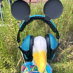 May include: A white goose wearing blue glitter headphones with black Mickey Mouse ears. The goose is wearing a blue and purple floral patterned shirt.
