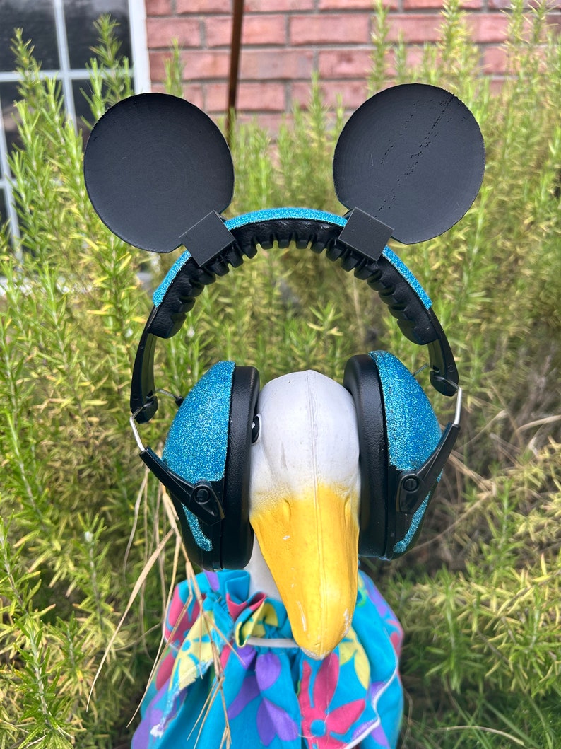 May include: A white goose wearing blue glitter headphones with black Mickey Mouse ears. The goose is wearing a blue and purple floral patterned shirt.