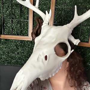 May include: A white deer skull mask with large antlers. The mask has eye and nose openings. The antlers are a bright white color. The skull is a matte white color. The mask is a costume or decorative item.