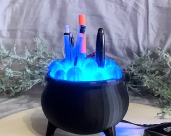 May include: A black cauldron-shaped desk organizer with a blue light emanating from the bottom. The cauldron is filled with blue and white marbles and has three pens sticking out of the top.