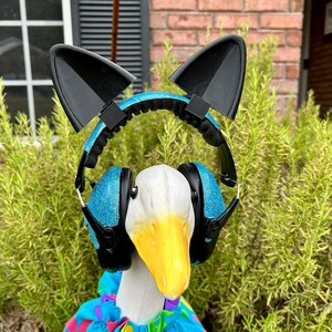 May include: A white goose wearing a blue and black headband with cat ears and blue glitter earmuffs. The goose is wearing a colorful floral print shirt.
