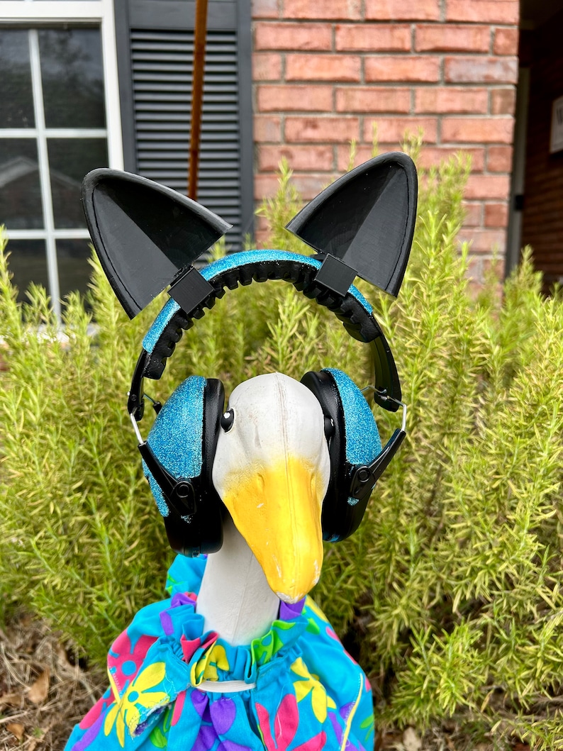 May include: A white goose wearing a blue and black headband with cat ears and blue glitter earmuffs. The goose is wearing a colorful floral print shirt.