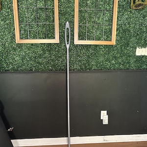 May include: A large, silver-colored sewing needle featuring a long, slender body and a prominent, elongated eye at the top. The needle has a smooth, metallic finish and tapers to a sharp point at the bottom, standing vertically against a wall.