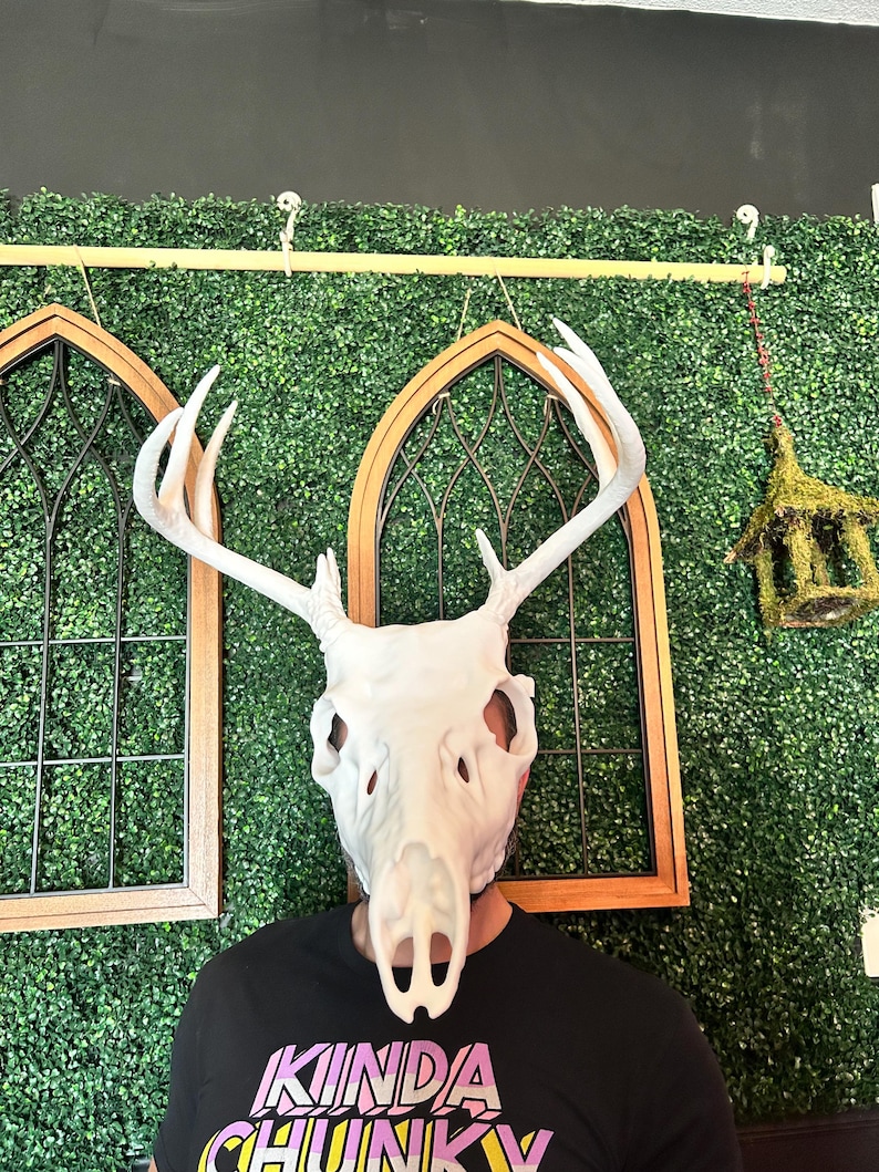 May include: A white deer skull mask with large antlers. The mask is worn over a person's face. The person is wearing a black t-shirt with the text "KINDA CHUNKY" in pink, yellow, and purple. The background includes decorative wooden framed windows and a green hedge.
