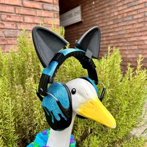 May include: A white goose wearing a blue and black pair of headphones with cat ears. The goose is wearing a blue and pink patterned shirt.