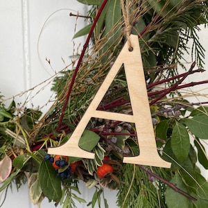 May include: A wooden letter A ornament, painted in a natural wood colour, hanging from a wreath of greenery.