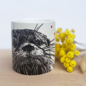 May include: A white ceramic mug with a black and white otter illustration. The mug has a cylindrical shape and red heart details. Yellow flowers are in the background. The mug is on a wooden surface.