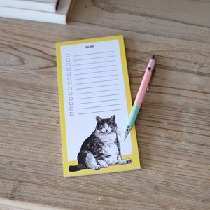 May include: A yellow notepad with a to-do list and a black and white illustration of a cat. The notepad has a pink pencil on top of it.