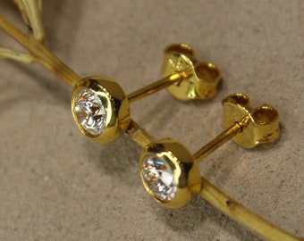Könnte beinhalten: Ein Paar goldene Ohrstecker mit klaren, runden Edelsteinen. Die Ohrringe sind in einer goldenen Lünettenfassung gefasst. Sie werden auf einem goldfarbenen Stiel mit einem getrockneten Blatt präsentiert.