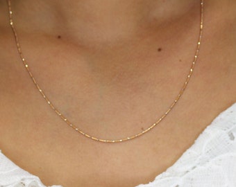 May include: A delicate gold necklace with small, evenly spaced beads. The necklace is worn against a white lace top. The gold chain is thin and appears to be a simple, elegant design.