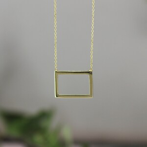 Op de afbeelding: Een delicate goudkleurige ketting met een rechthoekige hanger. De hanger is een eenvoudig, open frame-ontwerp dat aan een fijne ketting hangt. De achtergrond is wazig, met hints van groen gebladerte.