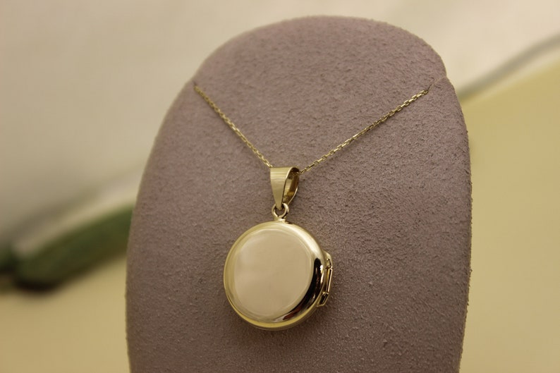 Puede incluir: Un collar con un colgante de medall&oacute;n redondo dorado. El colgante tiene forma circular pulida con una apertura con bisagras. La cadena es delicada y tambi&eacute;n dorada. El medall&oacute;n se exhibe sobre una superficie gris texturizada.