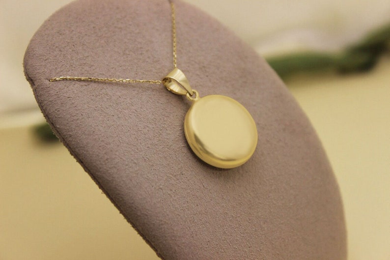 Puede incluir: Un collar con colgante dorado. El colgante es un disco liso y circular. La cadena del collar es fina y tambi&eacute;n dorada. La joya se exhibe sobre una superficie gris y texturizada.