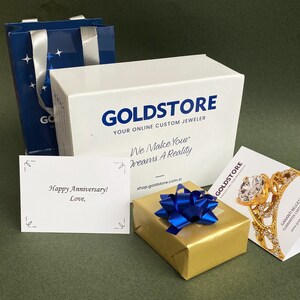Puede incluir: Un juego de regalo Goldstore, que incluye una caja blanca y una bolsa de compras azul. Un regalo envuelto en oro est&aacute; atado con un lazo azul, y una tarjeta dice "Happy Anniversary! Love,". Tambi&eacute;n es visible un anillo de oro.