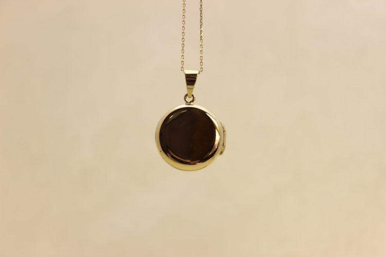 Puede incluir: Un collar con un medall&oacute;n dorado redondo. El colgante es de color oro brillante y reflectante, colgado de una delicada cadena. El medall&oacute;n est&aacute; cerrado, sobre un fondo neutro.