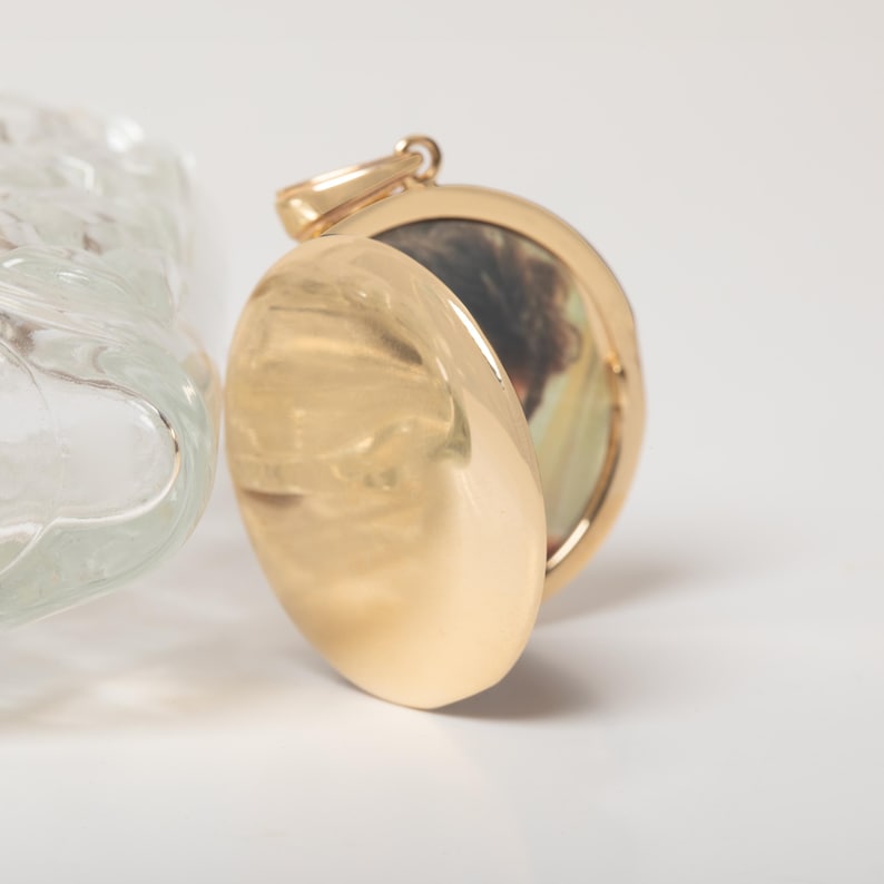 Puede incluir: Un colgante de medall&oacute;n dorado, parcialmente abierto para revelar una peque&ntilde;a imagen. El medall&oacute;n tiene una superficie lisa y reflectante y un lazo para una cadena. Fondo blanco.