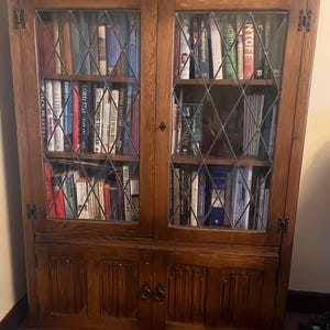 May include: A large, wooden bookcase with glass-paned doors and diamond-patterned metal grids. The bookcase is filled with books of various sizes and colors. The lower section has two doors with decorative carvings and ring pulls. The wood has a warm, brown finish.