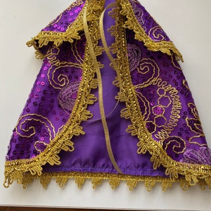 Puede incluir: Capa morada con ribete dorado y lentejuelas. La capa tiene un cierre de corbata y está decorada con un patrón floral.