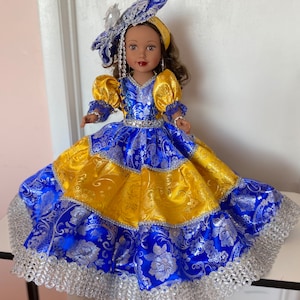 Op de afbeelding: Een pop in een blauw en gele jurk met zilveren bloempatronen en afwerking. De jurk heeft een volle rok en pofmouwen. De pop heeft bruin haar en draagt een bijpassende blauw en zilveren hoofdtooi.
