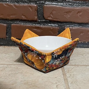 Può includere: Un copri ciotola in tessuto con un motivo floreale e di zucche. Il copri ciotola è piegato per contenere una ciotola in ceramica bianca.