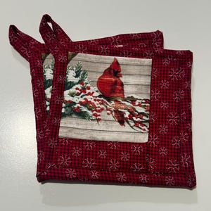 May include: Two red and black plaid potholders with a winter scene featuring a red cardinal bird perched on a snow-covered branch. The potholders have a square shape with a loop for hanging.