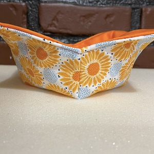 May include: A fabric bowl with a sunflower pattern in shades of yellow, orange, and white. The bowl is lined with orange fabric and has a white background.