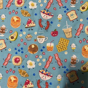 Op de afbeelding: Een lichtblauwe stof met een herhalend patroon van antropomorfe ontbijtproducten, waaronder spek, pannenkoeken, eieren, wafels, koffiekopjes, avocado's, croissants, muffins en fruit, allemaal met lachende gezichtjes en dunne zwarte ledematen.