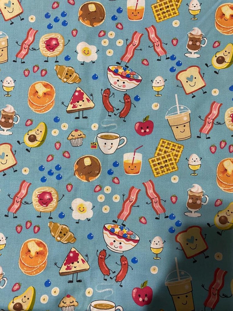 Op de afbeelding: Een lichtblauwe stof met een herhalend patroon van antropomorfe ontbijtproducten, waaronder spek, pannenkoeken, eieren, wafels, koffiekopjes, avocado's, croissants, muffins en fruit, allemaal met lachende gezichtjes en dunne zwarte ledematen.
