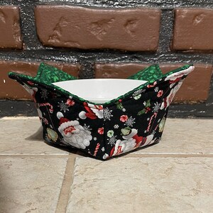 May include: A black and green quilted bowl cozy with a Santa Claus print. The cozy is holding a white ceramic bowl.