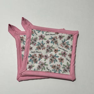 Puede incluir: Dos agarraderas de olla de color rosa y blanco con un patrón floral y el texto "Apple Blossom" repetido en todo.