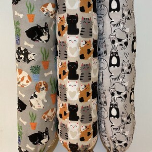 May include: Three fabric bag dispensers with different cat and dog patterns. The dispensers are grey with white and black designs. The dispenser on the left has a dog pattern, the middle dispenser has a cat pattern, and the right dispenser has a cat pattern.