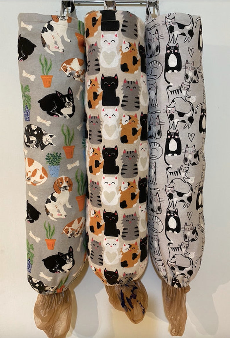 May include: Three fabric bag dispensers with different cat and dog patterns. The dispensers are grey with white and black designs. The dispenser on the left has a dog pattern, the middle dispenser has a cat pattern, and the right dispenser has a cat pattern.