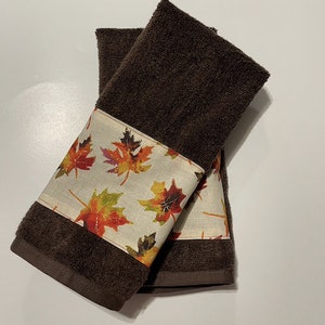 May include: Two brown kitchen towels with a white band featuring a colourful autumn leaf pattern.