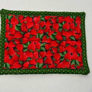May include: Rectangular quilted placemat featuring a vibrant pattern of red strawberries with green leaves. The border is a dark green fabric with white polka dots. The placemat is likely used for dining or kitchen decor.