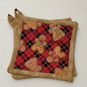 May include: Two square quilted pot holders. The center fabric features a red and black plaid pattern with gingerbread men. The edges are trimmed with a tan fabric with a small leaf pattern. Each pot holder has a loop for hanging.
