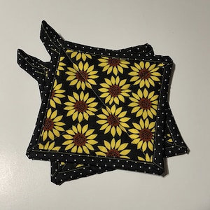May include: Two square pot holders with a black background and a pattern of yellow sunflowers with brown centers. The edges are black with white polka dots. The pot holders are quilted.