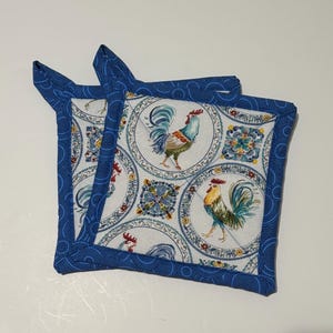 May include: Two square pot holders with a rooster and floral design. The pot holders have a white background with a repeating pattern of roosters and floral medallions. The edges are trimmed with a blue fabric.