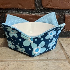 May include: A blue and white floral bowl cozy with a white bowl inside. The cozy is made of fabric with a blue background and white flowers with blue centers.