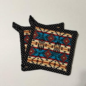 May include: Two square fabric pot holders with a Native American design. The center features a turquoise, red, and tan geometric pattern with dream catchers. The edges are black with white polka dots. The pot holders have a small loop for hanging.