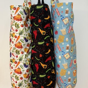 Op de afbeelding: Drie cilindervormige stoffen zakken voor het opbergen van plastic tasjes, elk met een unieke print. De linkerzak is wit met kleurrijke groenten, de middelste zak is zwart met diverse pepers en de rechterzak is lichtblauw met ontbijticoontjes.