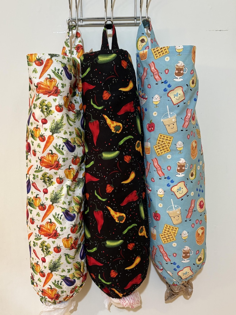 Op de afbeelding: Drie cilindervormige stoffen zakken voor het opbergen van plastic tasjes, elk met een unieke print. De linkerzak is wit met kleurrijke groenten, de middelste zak is zwart met diverse pepers en de rechterzak is lichtblauw met ontbijticoontjes.