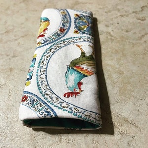 May include: A white fabric utensil holder with a teal lining. The fabric has a repeating pattern of blue, green, and yellow roosters in circular frames.