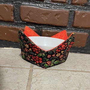 May include: A black and orange fabric bowl cozy with a floral pattern. The cozy is folded and a white bowl is inside.