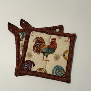 Peut inclure: Deux maniques matelassées brunes avec un motif de coq. Les maniques ont une bordure brune et un fond crème avec un motif de coq et de fleurs.