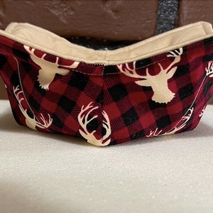 Puede incluir: Un cuenco de tela con un estampado de cuadros de búfalo rojo y negro, con siluetas de ciervos blancos. El interior es de color beige liso. El cuenco está diseñado para contener alimentos.