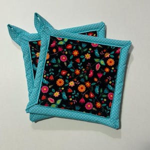 Puede incluir: Un par de agarraderas de olla azul turquesa con lunares blancos. Las agarraderas están forradas con una tela negra con un estampado floral y de pájaros coloridos.