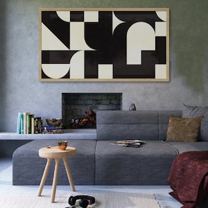 May include: A framed abstract art print with a geometric design in black and white hangs above a gray sectional sofa. The artwork features shapes like squares, semi-circles, and triangles. A small wooden stool and headphones are in the foreground.