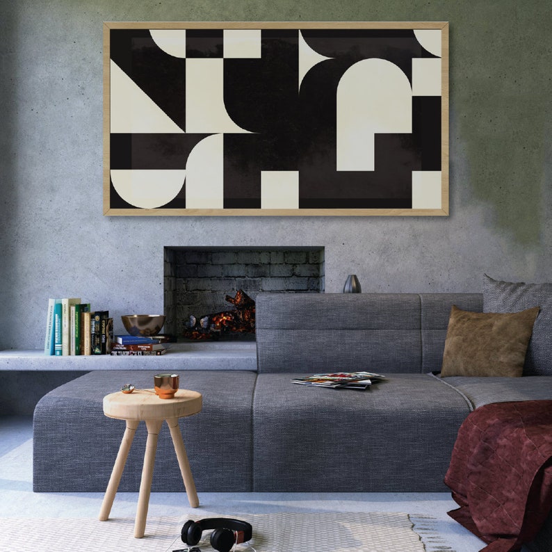 May include: A framed abstract art print with a geometric design in black and white hangs above a gray sectional sofa. The artwork features shapes like squares, semi-circles, and triangles. A small wooden stool and headphones are in the foreground.