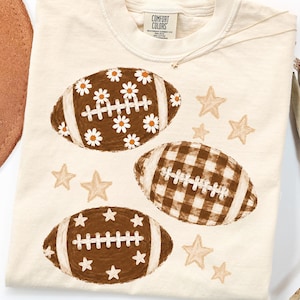 May include: A cream-colored t-shirt featuring a graphic print of three footballs and several stars. One football has a white daisy pattern, one has a brown and white gingham check pattern, and one is solid brown with white star accents.