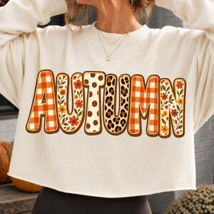 Op de afbeelding: Een crèmekleurig, langmouwig cropped sweatshirt met het woord AUTUMN op de borst gedrukt. Elke letter heeft een uniek patroon, waaronder oranje gingham, bloemenprints, bruine stippen en een luipaardprintontwerp.